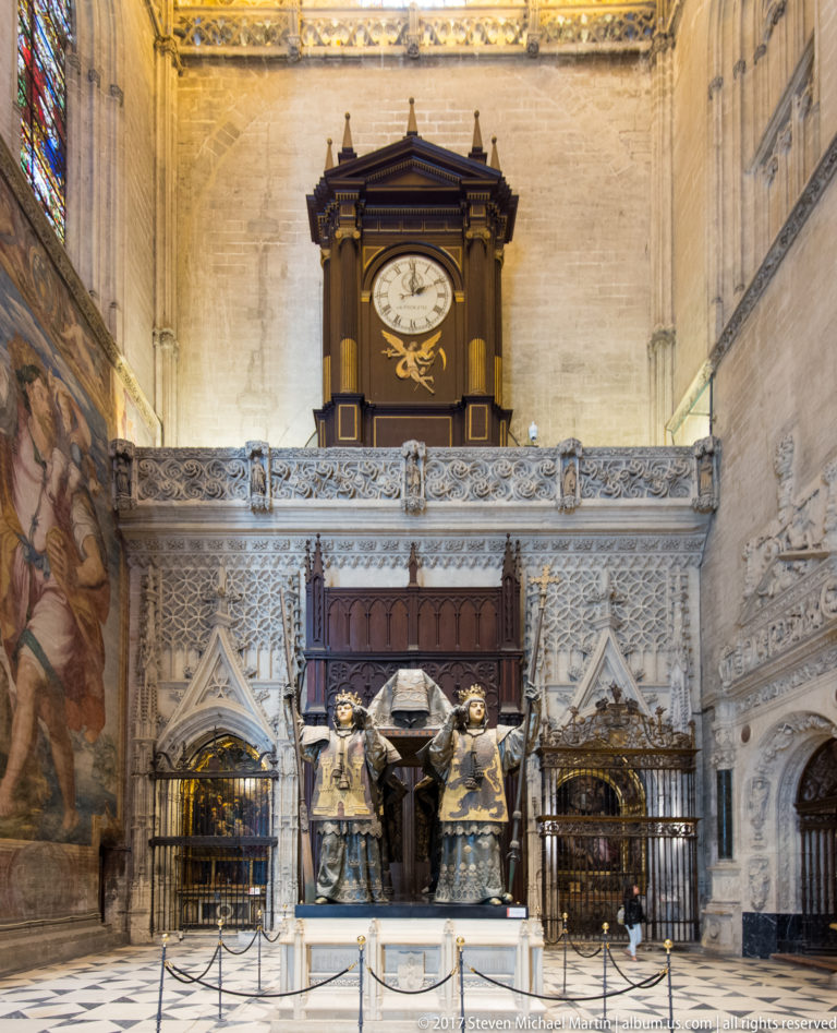 Wide cathedral view of Columbus’s tomb below a clock, with carved stonework, grilles, and a large wall painting.
