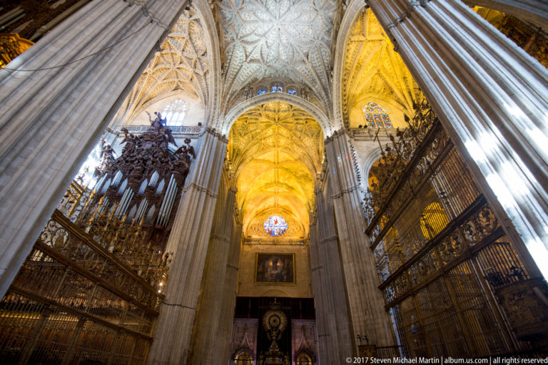 Angled interior view of Seville Cathedral with massive pier, carved wooden screens, iron grilles, and illuminated ribbed vaults.