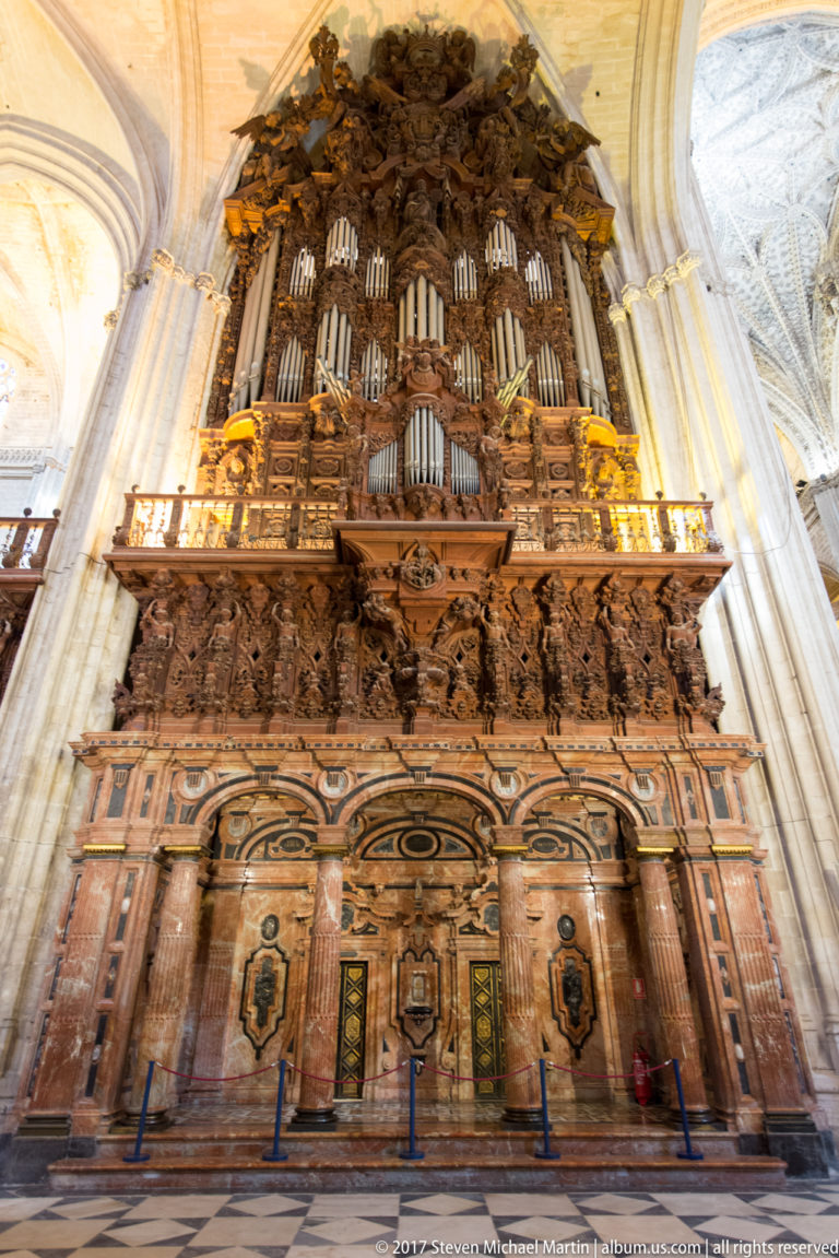 Large cathedral pipe organ with many metal pipes in an elaborate dark carved wooden case above paneled woodwork.