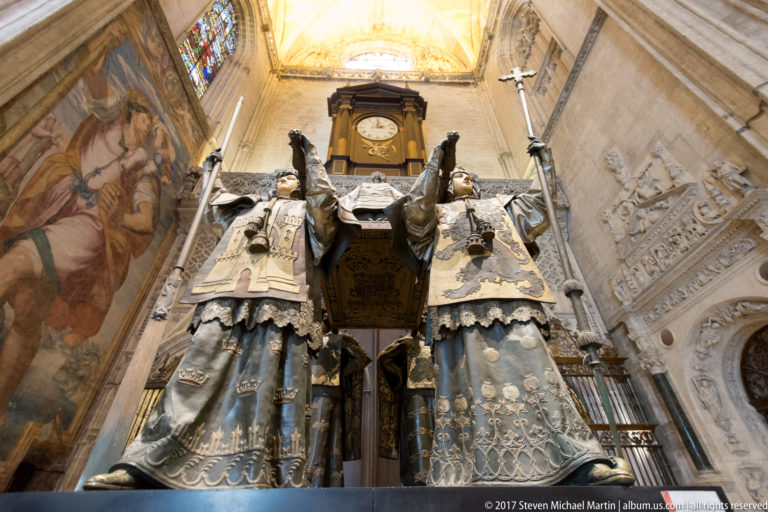 Low-angle close view of two crowned figures supporting Columbus’s sarcophagus, with a clock and cathedral wall behind.