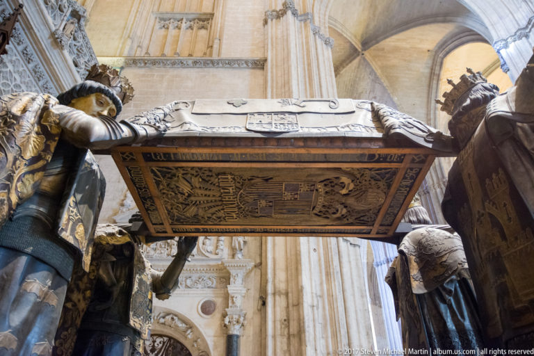 Low view beneath Columbus’s decorated sarcophagus as crowned figures in patterned robes support it at the corners.