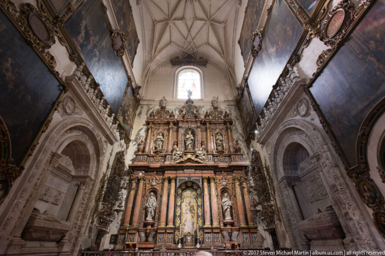 Tall ornate altarpiece at the end of a vaulted cathedral chapel, with side arches, dark paintings, and high window.