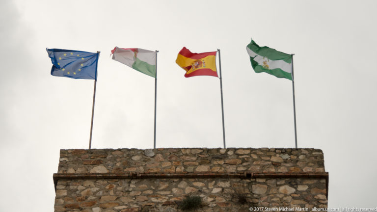 European Union, local, Spanish, and Andalusian flags flying above a rough stone parapet.
