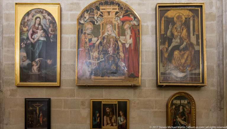 Several framed and panel religious paintings of different sizes arranged on a pale stone wall inside Seville Cathedral.