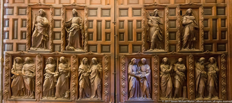 Rows of carved standing religious figures arranged in two tiers against warm wooden paneling inside Seville Cathedral.