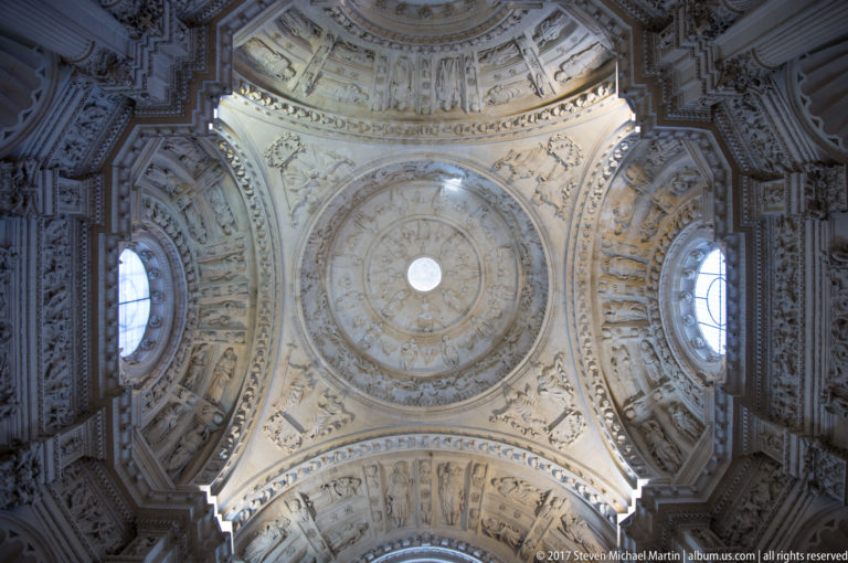 Upward view of an ornate pale dome with coffers, sculpted reliefs, pendentives, cornices, and arched windows.