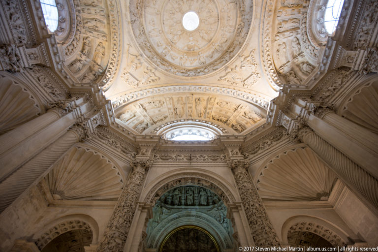 Ornate pale domed ceiling above shell-vaulted arches, relief panels, pilasters, and a green-toned feature beyond.