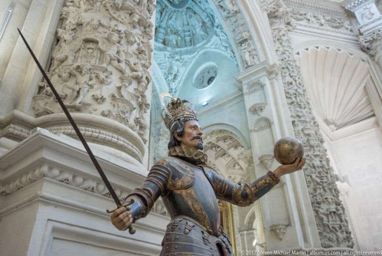 Armored moustached figure with crown-like headgear holding a long sword and orb beneath richly carved pale stonework.