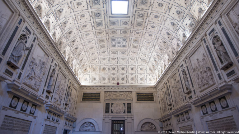 Long pale chamber with patterned coffered barrel vault, sculpted wall figures, relief panels, and doorway at far end.