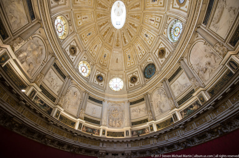 Wide upward view of an ornate coffered dome, stained-glass windows, painted roundels, reliefs, pilasters, and decorated drum.