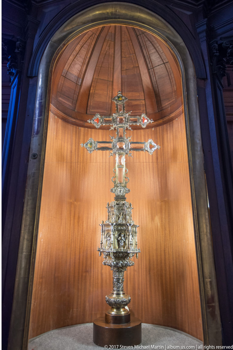 Tall ornate silver processional cross with sculpted details and elaborate base displayed upright in a warm-lit niche.