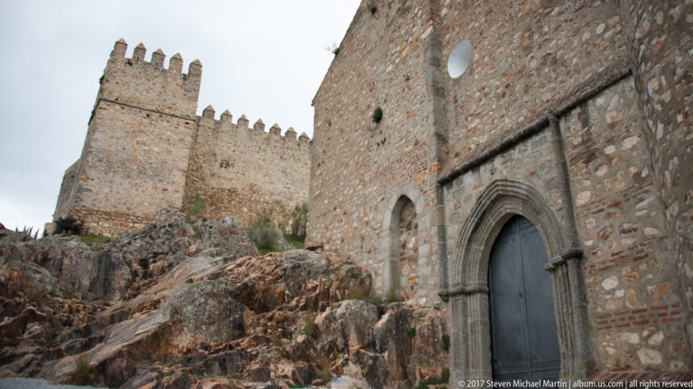 Pointed-arch doorway in rough stone masonry below crenellated walls and a square fortified tower.