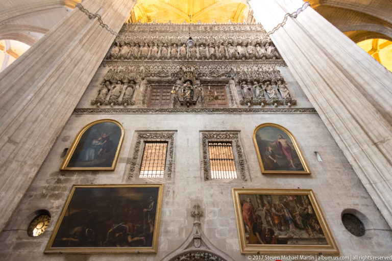 Interior cathedral wall with framed paintings, carved stone frieze, barred windows, and massive columns rising on both sides.