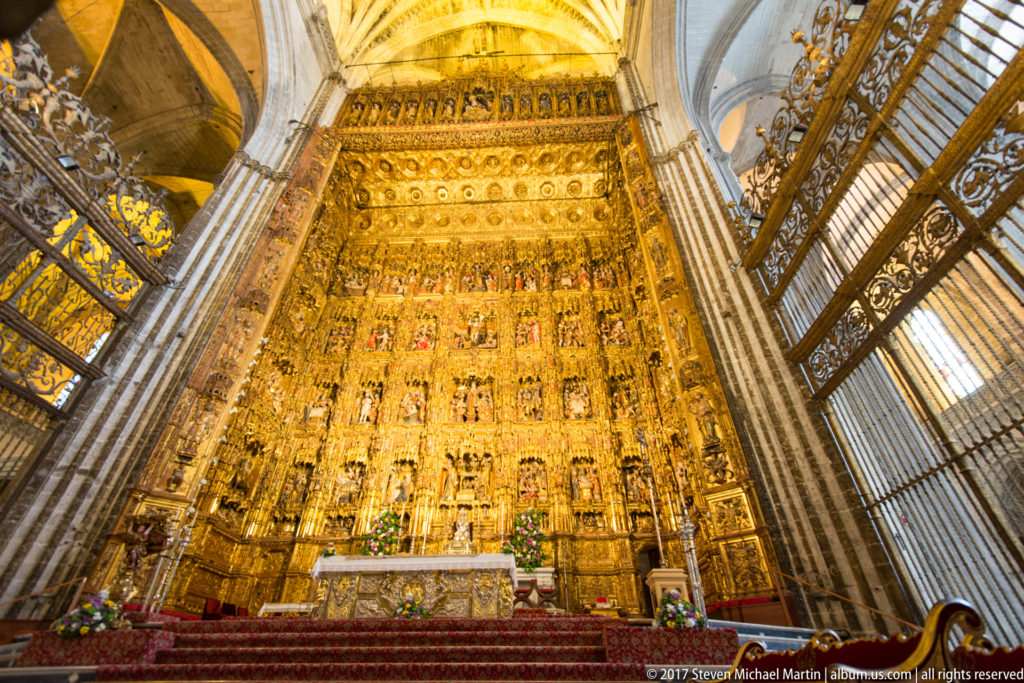 Gilded monumental altarpiece rising behind the altar, framed by tall cathedral columns and metal choir screens.