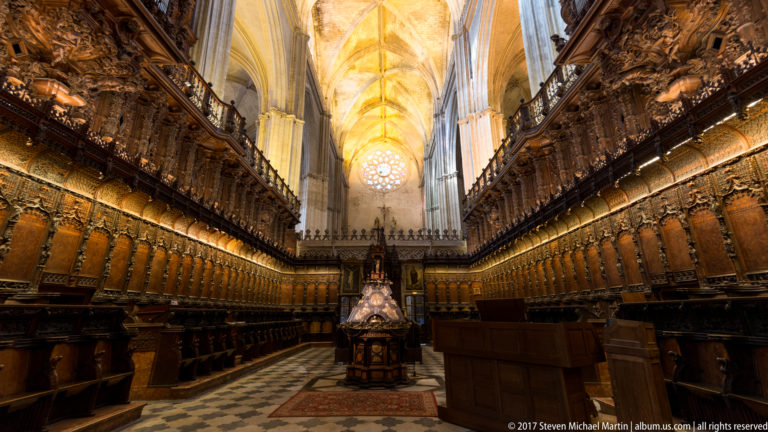 Long cathedral choir lined with dark carved wooden stalls beneath a high vault, with stained glass glowing at the far end.