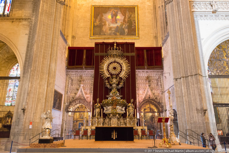 Large ornate silver-gilt monstrance with a radiant circular center displayed on a platform against red drapery inside the cathedral.