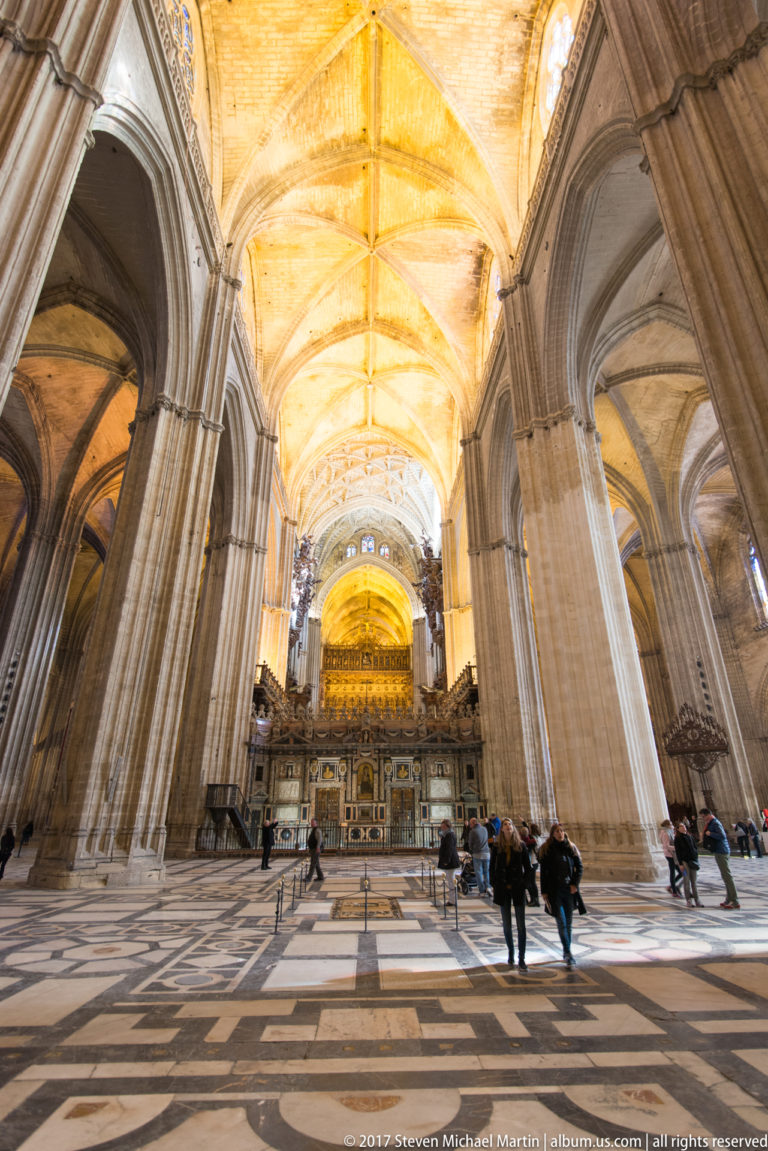 Wide cathedral interior with massive stone piers, high ribbed vaults, patterned floor, distant screen, and several visitors.