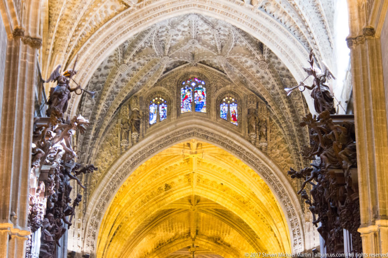 Ornately carved Gothic arch with tracery and three stained-glass windows above, flanked by dark wooden choir carving.