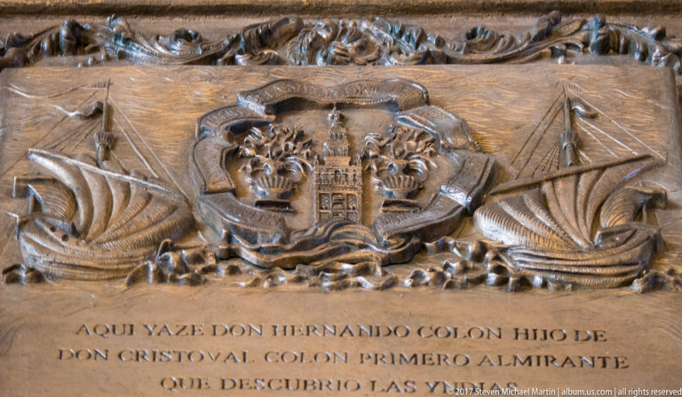 Carved stone tomb slab with Spanish inscription, central heraldic shield, and sailing ships on either side.
