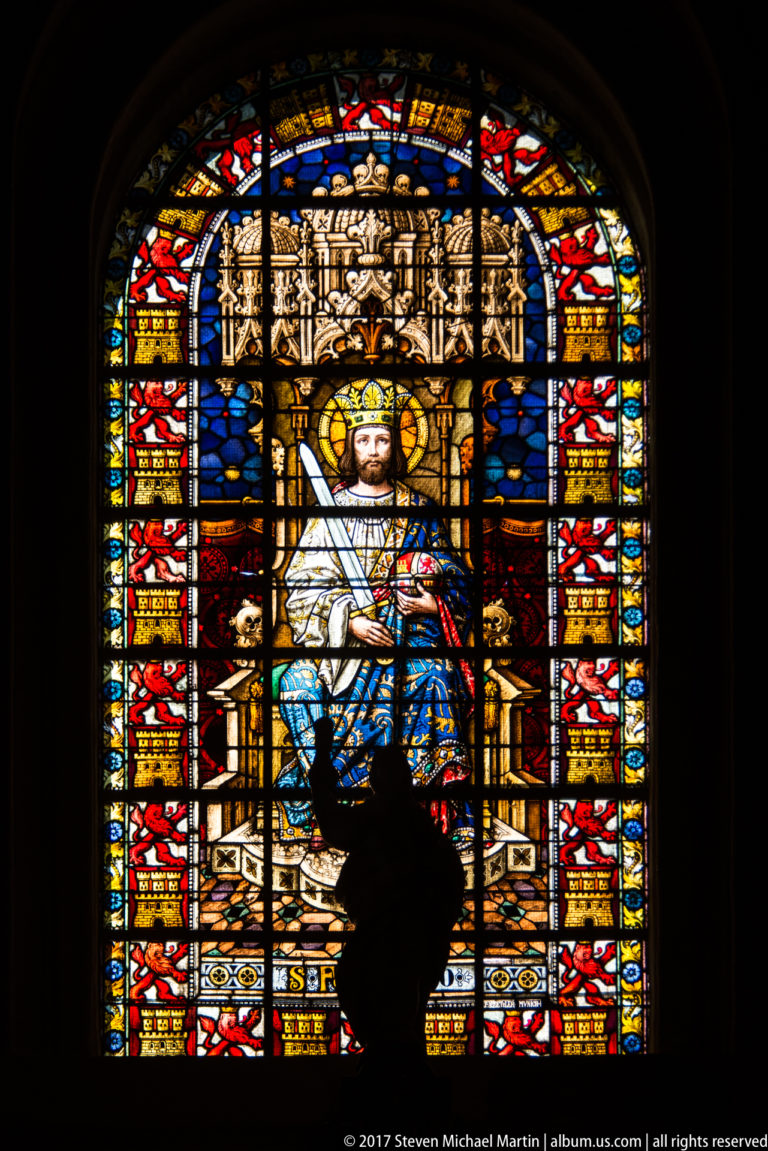 Colorful stained-glass window with an enthroned crowned figure, partly silhouetted by a visitor standing in front.