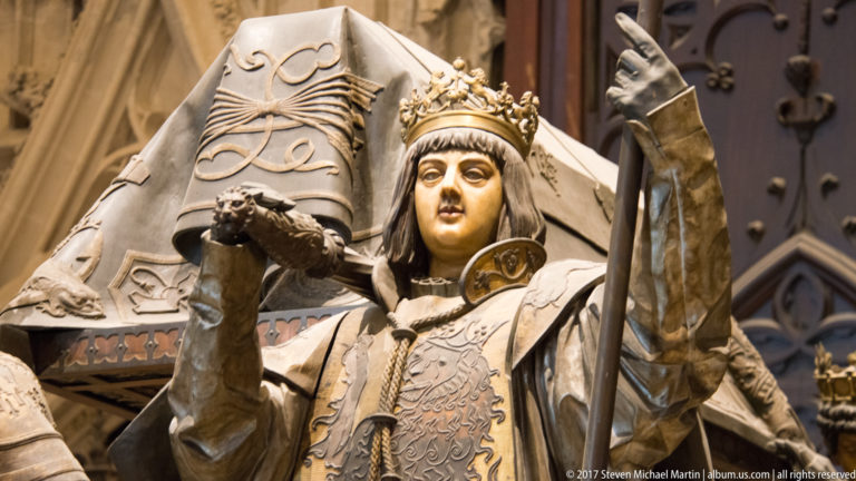 Close view of a crowned armored bearer figure from the tomb of Christopher Columbus, with heraldic shield behind.