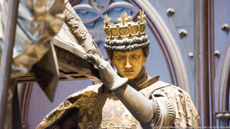 Crowned sculptural bearer with lowered gaze and raised hands supporting part of the Columbus tomb.
