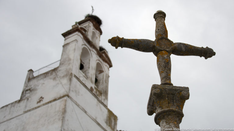 Weathered stone cross seen from below, with a white church bell gable blurred behind it.