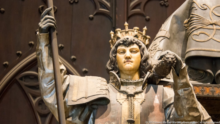 Frontal close-up of a crowned sculptural bearer holding a staff beside a large shield on the Columbus tomb.
