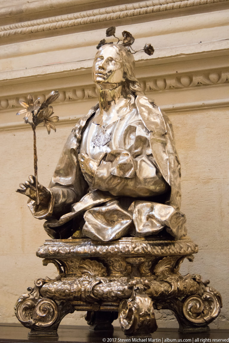 Seated silver-toned female saint holding a small cross on an ornate scrollwork base.