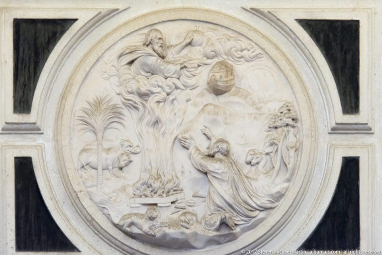 Circular pale stone relief of a kneeling figure facing a heavenly scene, set within a rectangular architectural panel.