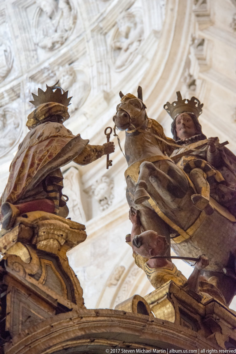 Low-angle view of three crowned polychrome religious figures in gilded garments beneath a pale church ceiling.