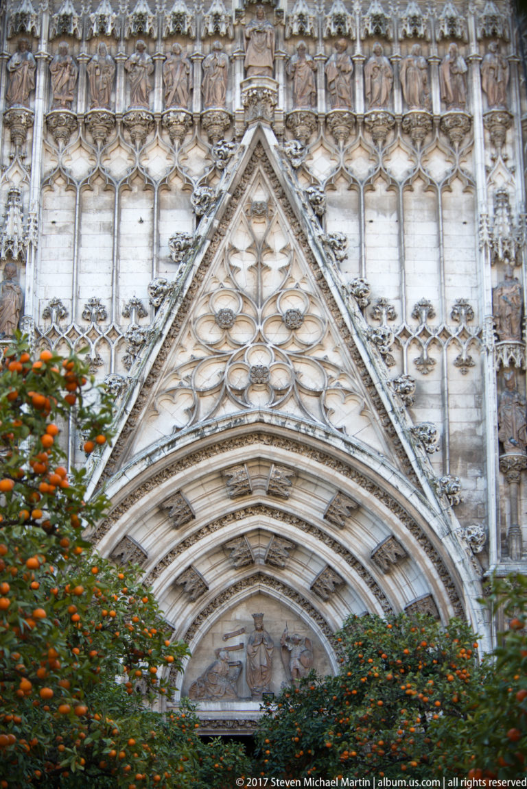 Pointed Gothic cathedral portal with layered carved arches and tracery, partly screened by orange trees.
