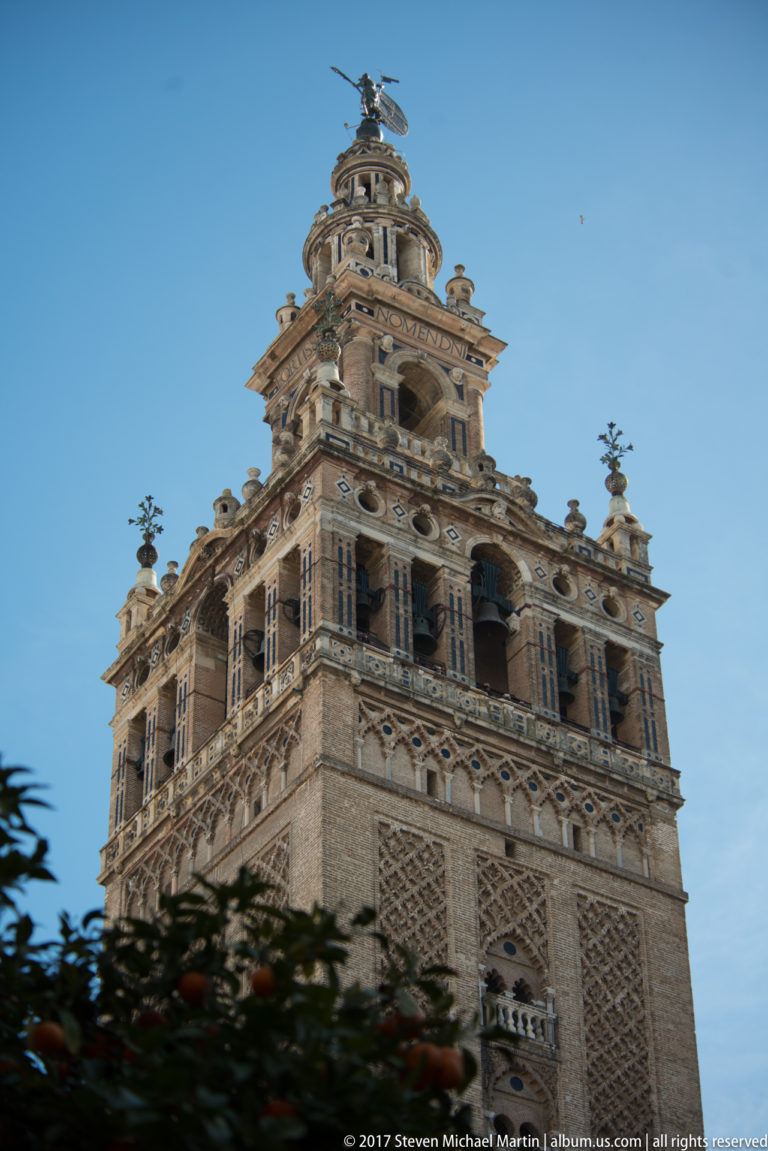 Upper portion of the Giralda with patterned masonry, Renaissance bell stages, and the summit weather vane against blue sky.