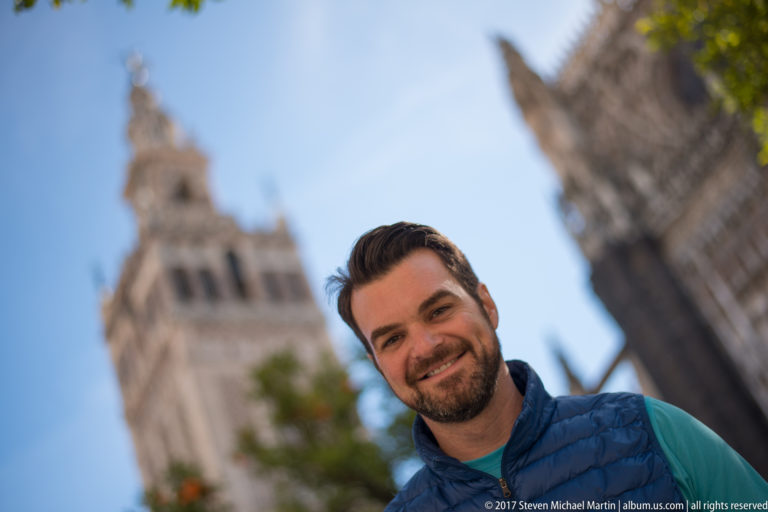 Smiling man in a blue jacket in sharp foreground with the Giralda and cathedral blurred behind him.
