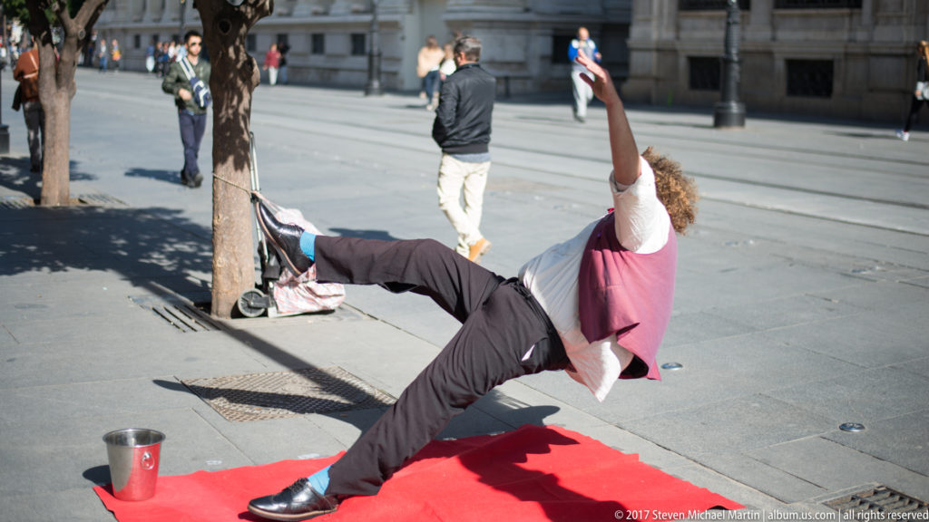 Street performer in black trousers and pink vest dancing on a red mat, with pedestrians in the plaza behind.