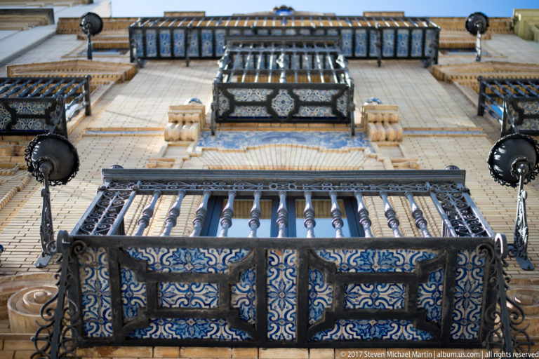 Upward view of stacked balconies with blue-and-white ceramic tiles, dark iron railings, and cream-colored masonry.