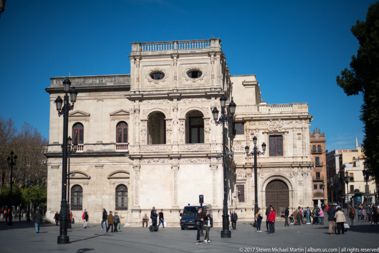 Pale carved façade of Seville City Hall fronting a plaza with pedestrians, lampposts, and vehicles.