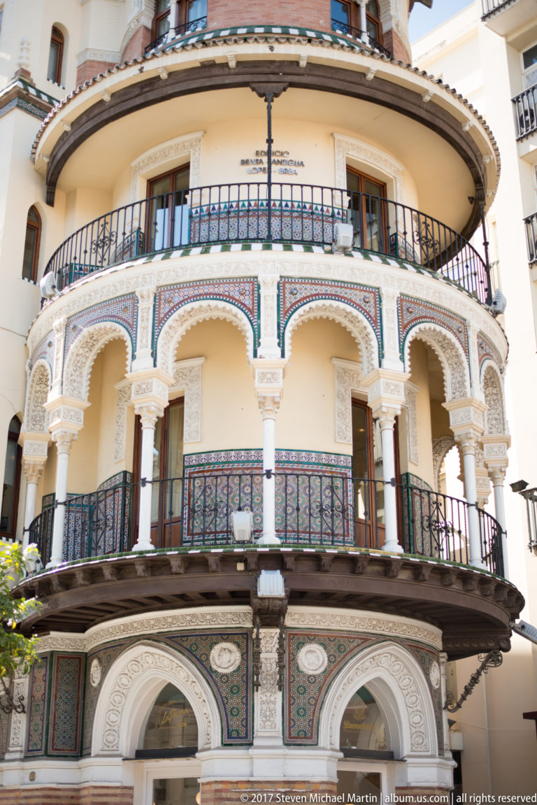 Rounded building corner with stacked arcaded balconies, patterned ceramic panels, and black wrought-iron railings.