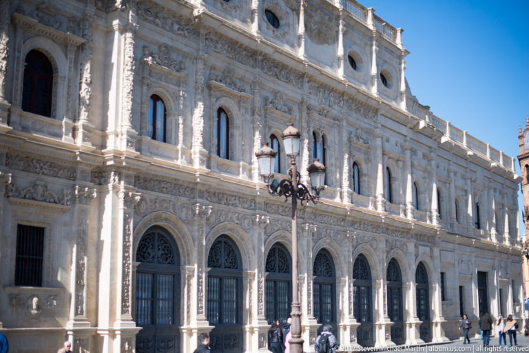 Oblique view along a highly carved pale-stone civic façade with arched windows, relief ornament, and decorative streetlamps.