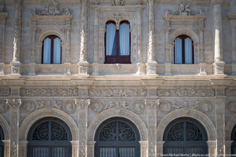 Close view of carved stone façade with upper windows, lower arched openings, relief bands, columns, and dark grilles.