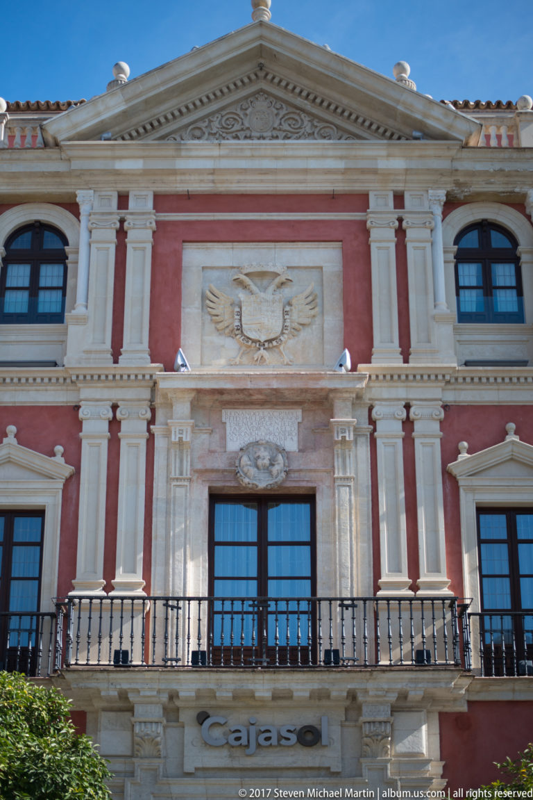 Red classical building façade with cream trim, central relief, triangular pediment, balconies, and visible Cajasol signage.