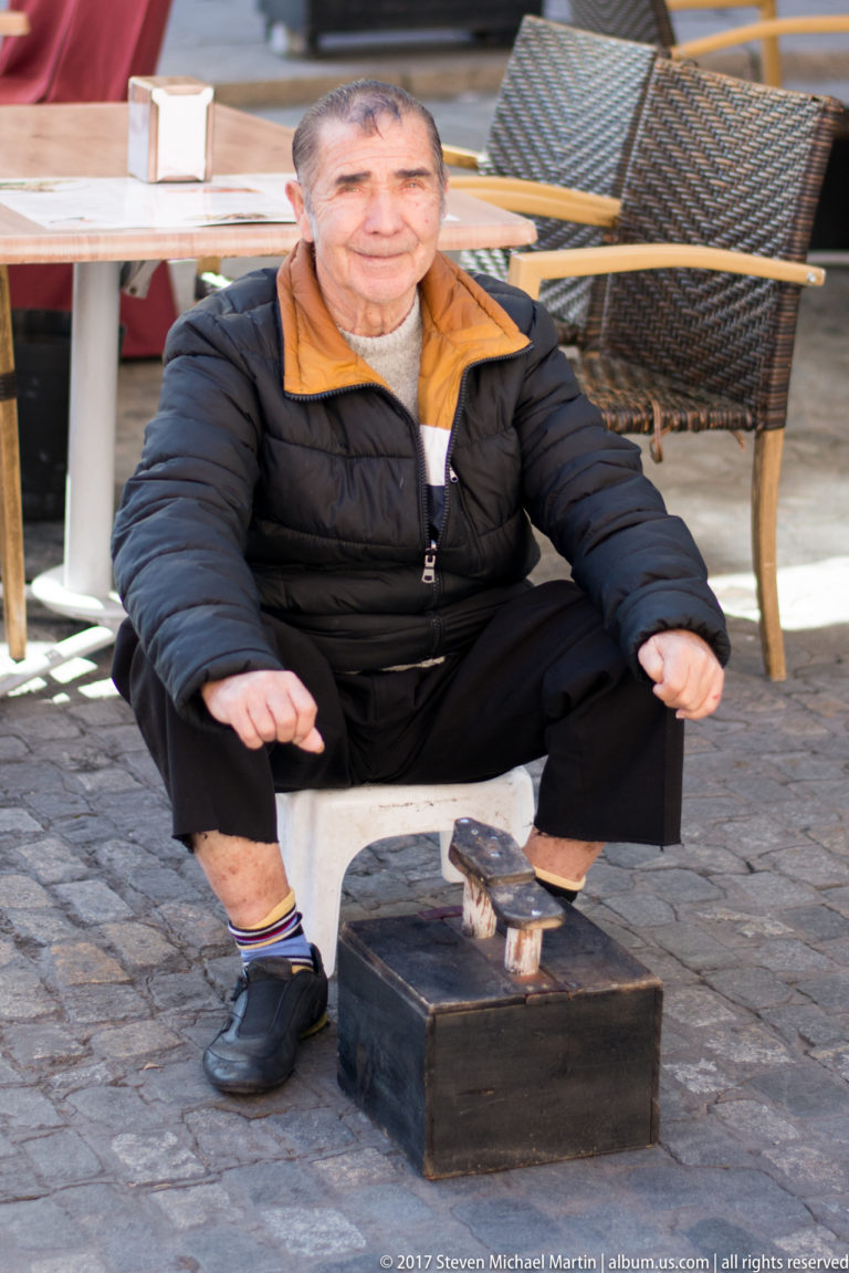 Seated street performer in dark clothing with a small puppet or figurine standing on a wooden box in front.