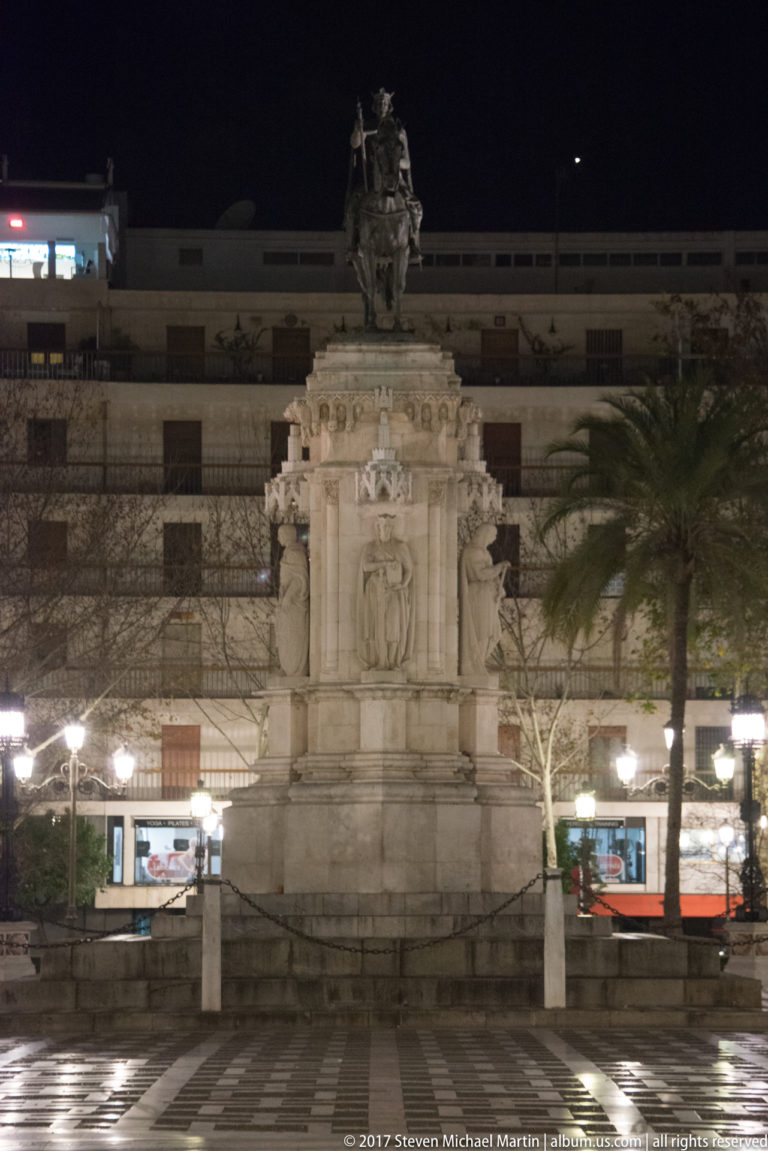 Illuminated stone monument with sculptural group and heraldic pedestal in a palm-lined city square at night.