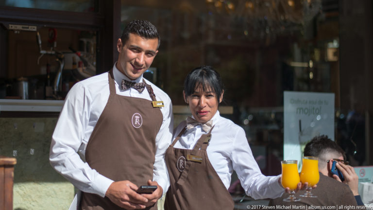 Two restaurant servers in white shirts and brown aprons stand outside a doorway, one holding two orange drinks.