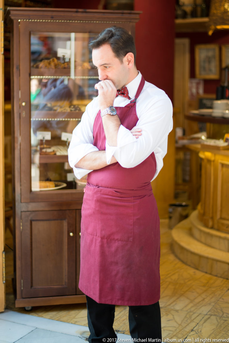 Waiter in a burgundy apron stands with folded arms inside a wood-paneled restaurant, holding a cloth to his face.