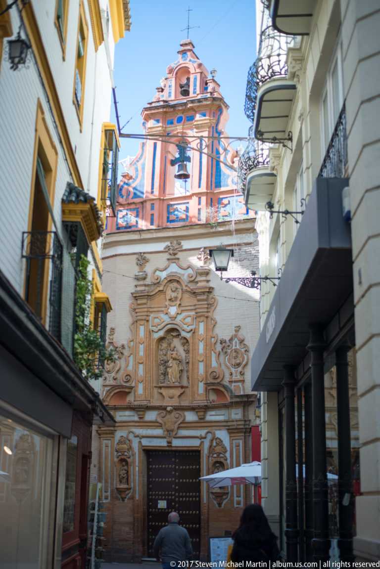 Ornate ocher-and-white Baroque church façade with sculpted portal and bell tower at the end of a narrow Seville street.