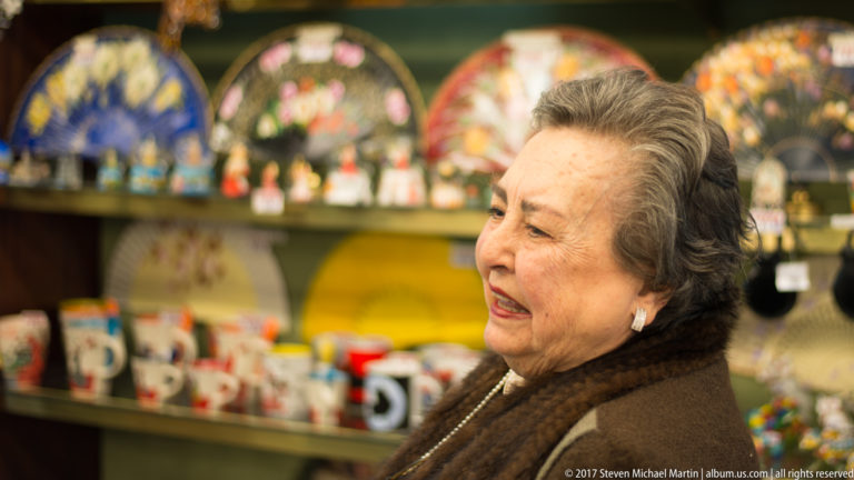 Older woman in profile inside a shop with colorful ceramics, fans, mugs, and souvenirs on shelves behind her.