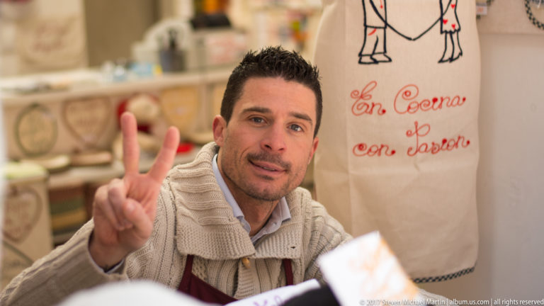 Man seated at a restaurant table raises two fingers toward the camera beside an embroidered “En Cocina con Javier” textile.