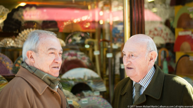 Two older men in coats converse outside a shop window displaying numerous decorative folding fans.