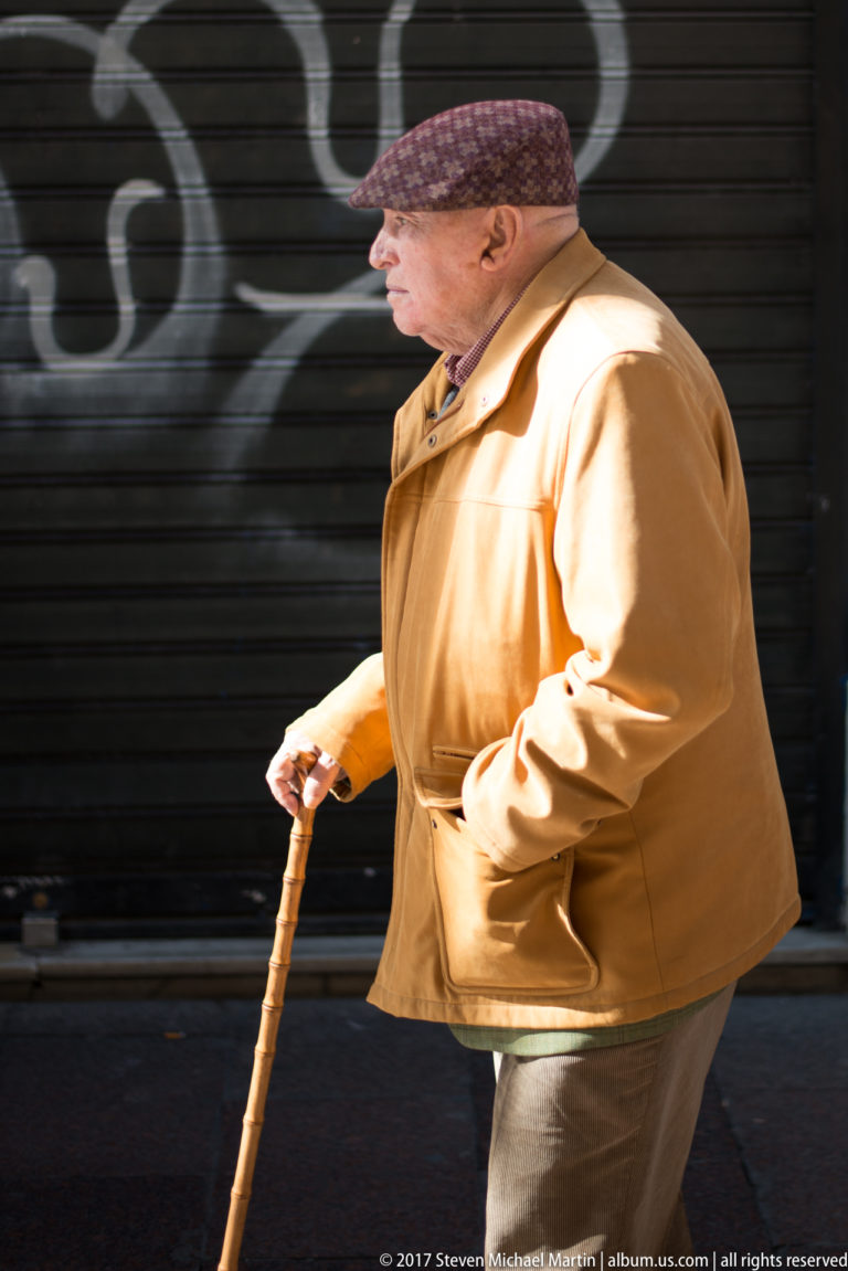 Older man in a tan jacket and flat cap walks with a wooden cane past a dark shuttered storefront.